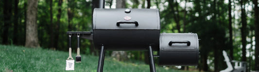 A Smokin' Joe barrel grill with attached offset smoker on the right and side shelf on the left. A spatula and a brush hang from the side shelf on the left.