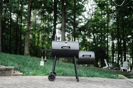 The Smokin' Pro barrel grill with offset smoker set up outside on a brick patio in front of a green lawn with blurry trees in the background.