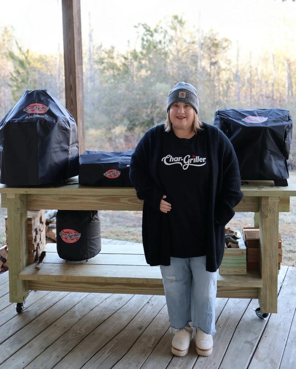 Jennifer Clark, wearing a Char-Griller t-shirt, stands in front of a rolling wooden table. On the table are Char-Griller grills: AKORN Jr., Flat Iron Portable 17-inch griddle, and Portable Side Fire Box.  