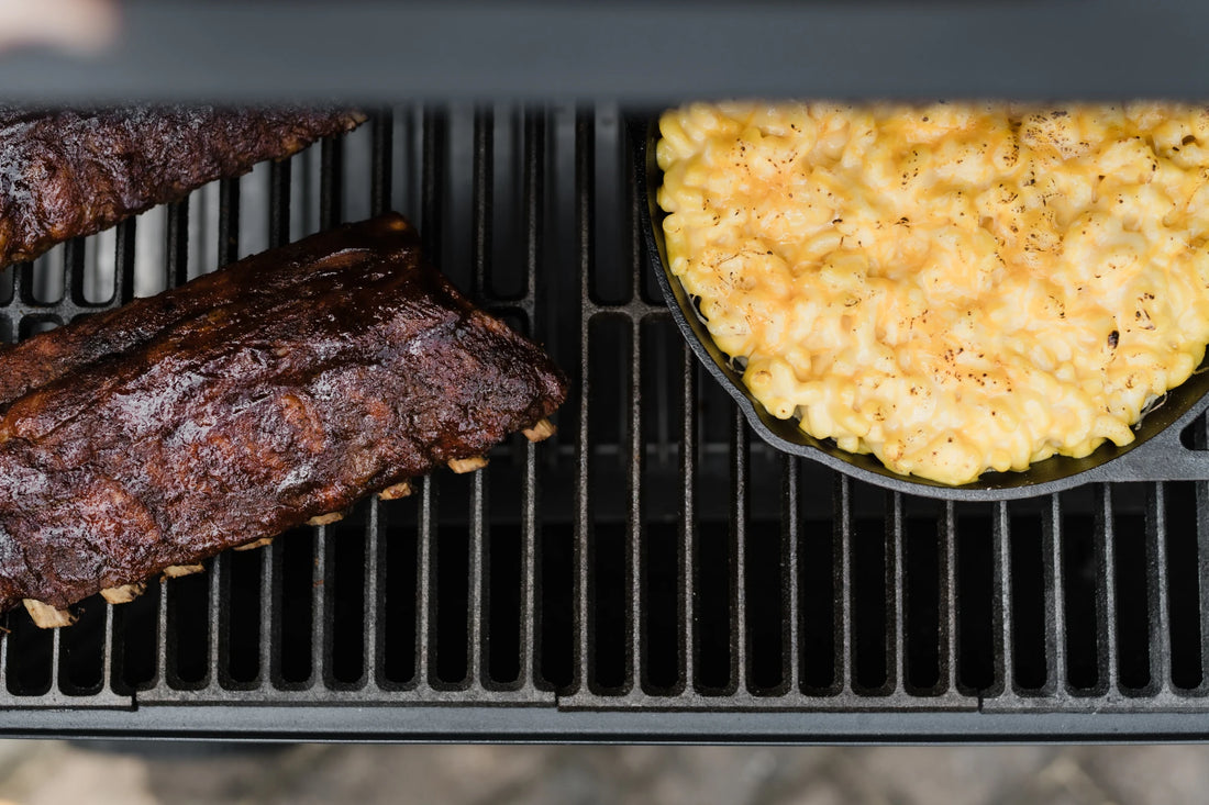 A cast iron skillet full of mac and cheese sits on a grill grate next to 2 sauce-coated racks of ribs