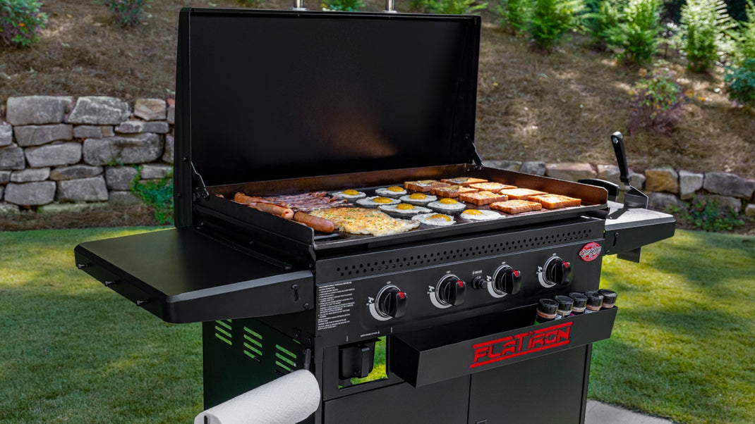 A 4-burner Flat Iron griddle sits on a paved patio in front of a landscaped lawn. The lid is open to show eggs, French toast, hashbrowns and more.