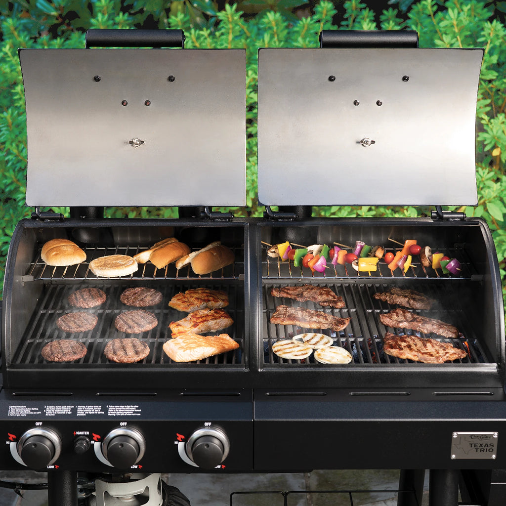 Texas Trio grill with both covers open, seen from above showing burgers, salmon fillets, steaks, and onion slices cooking on the main grates. Buns and vegetable skewers cook on the warming racks.
