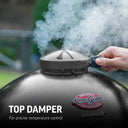 Smoke billows from the adjustable damper mounted at the very top of the grill dome as someone adjusts the setting. Text reads: top damper for precise temperature control.