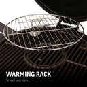 Closeup of the removable warming rack installed in an open AKORN grill. Circular rack has raised edges to stop food falling onto the cooking grate. Text reads: Warming rack to keep food warm.