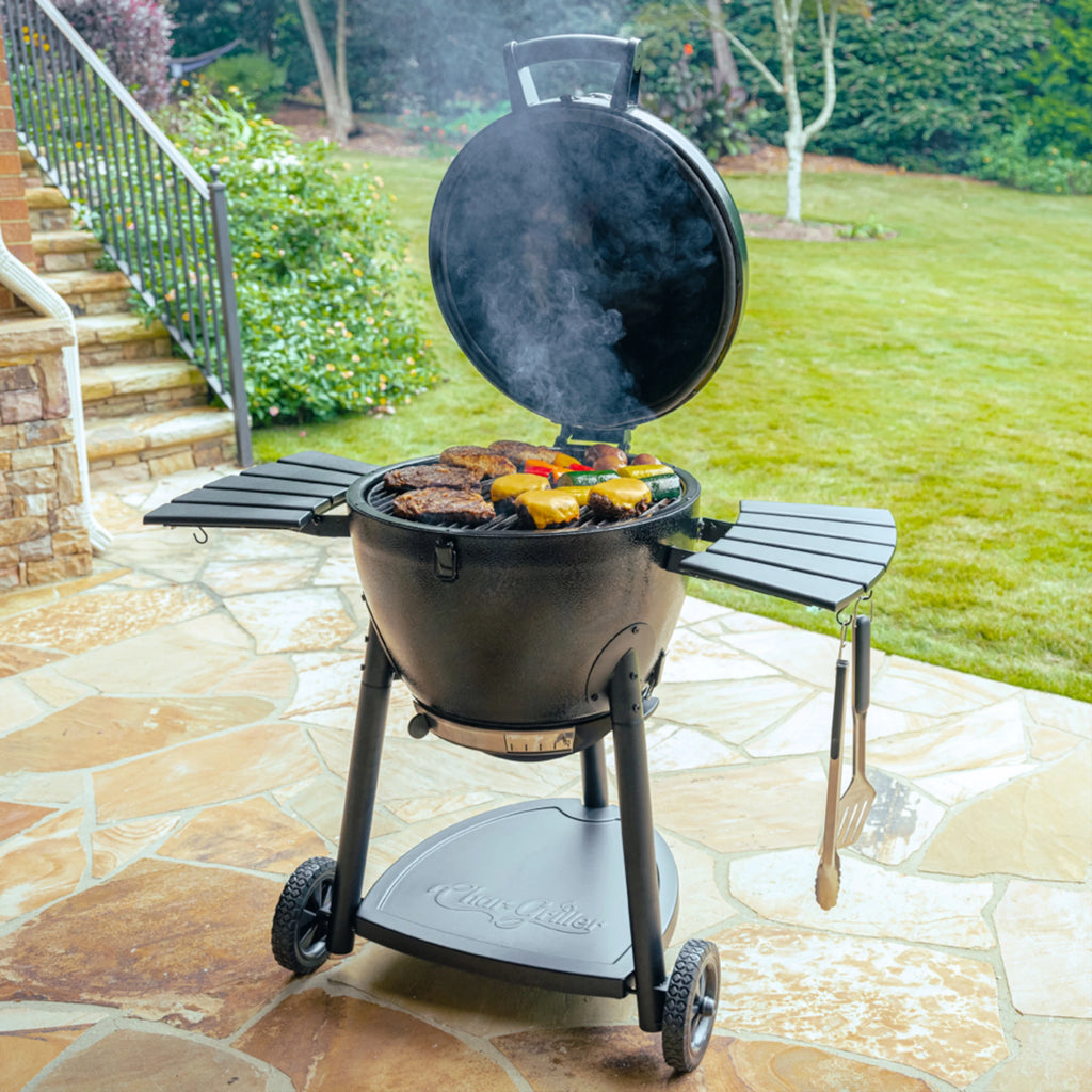 An open AKORN grill with a variety of food cooking on it. The grill is placed on a stone patio near the bottom of a flight of stone steps and in front of a green lawn with bushes in the background.