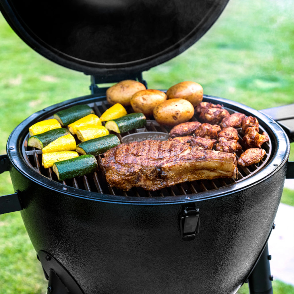 Closeup of meats and vegetables cooking on a black AKORN grill. The cooking grate has the Char-Griller logo in the center of it.