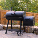 Dakota grill with side fire box installed on the right side. The grill sits on a paved outdoor patio in front of a wall made from small boulders in front of some bushes and a wooden privacy fence.