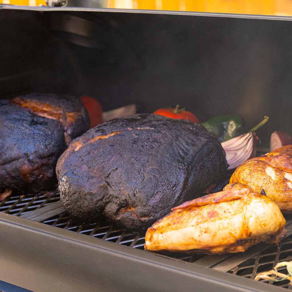 2 smoked pork butts with visible bark sit on the cooking grate of a Dakota grill next to chicken halves. Whole peppers and red onion wedges peek out from behind them.