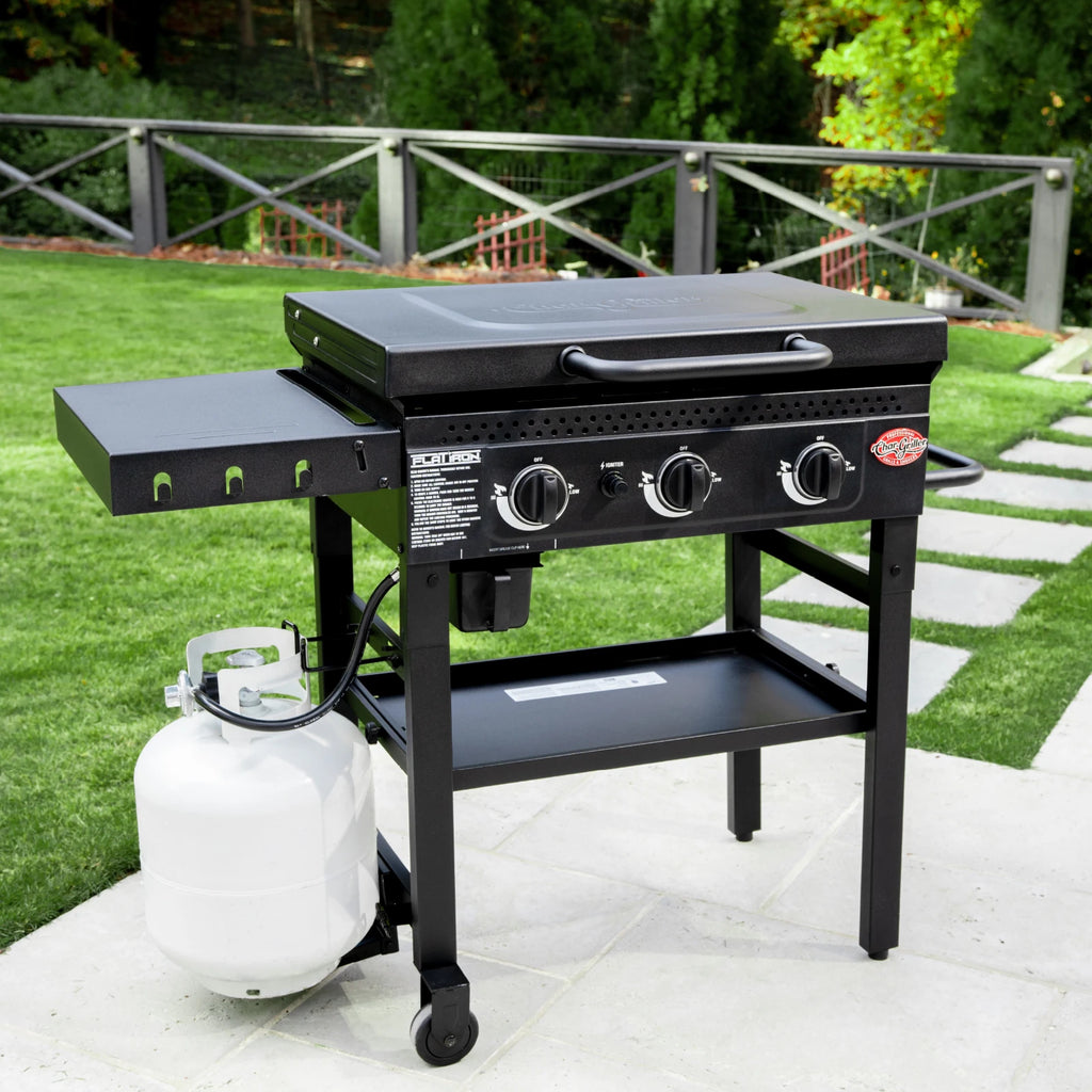 A 3-burner Flat Iron Griddle with lid and side shelf sitting closed on a cement patio in front of a walkway and metal fence