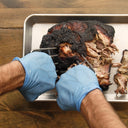 Someone wearing blue gloves uses 2 meat claws to pull apart a cooked butt
