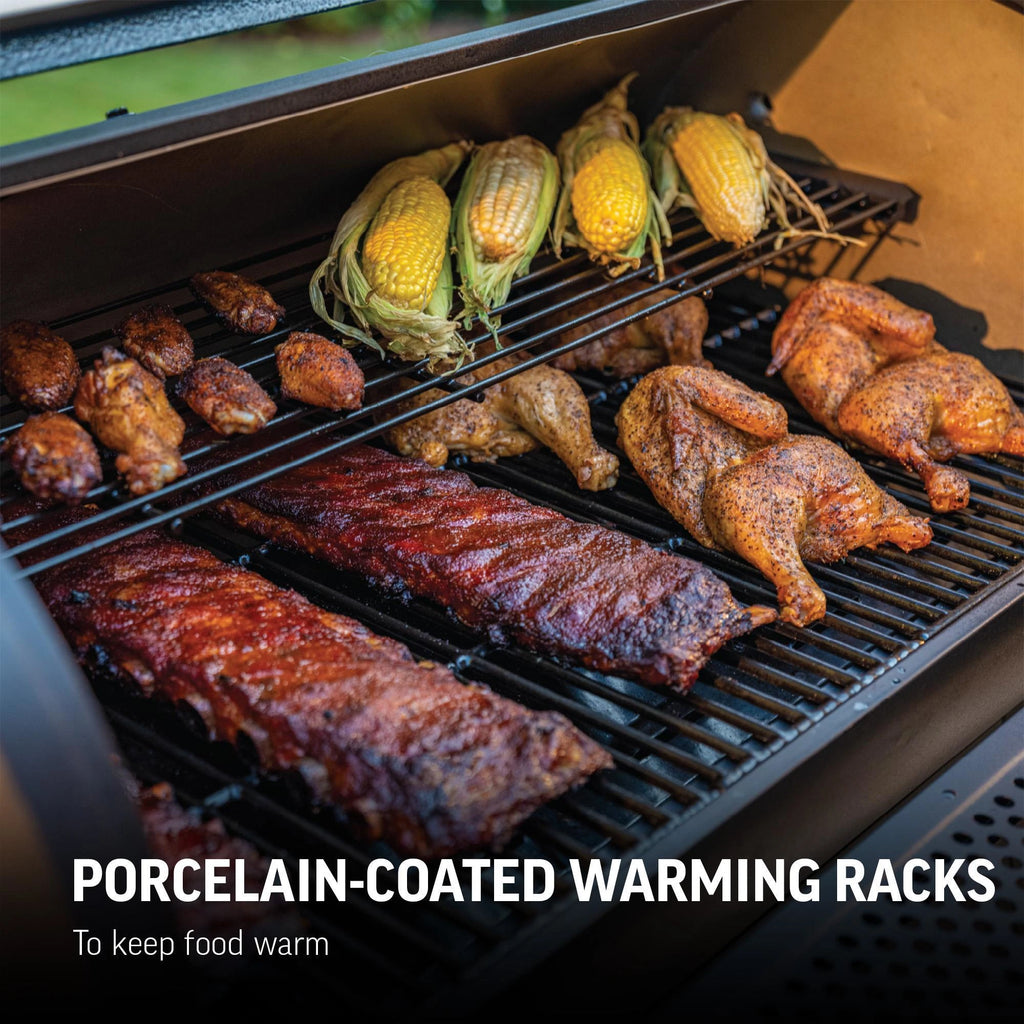 Ears of corn and chicken wings rest on the warming rack above chicken halves and racks of ribs on the main cooking grate. Text overlay reads: Porcelain-coated warming racks to keep food warm.