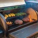 Steaks and veggies cook in an open Gravity Fed 980 grill. The perforated metal front shelf is in the upright position and runs the entire width of the grill body.