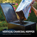 Person loading charcoal into a vertical charcoal hopper with text highlighting capacity and use. Text reads: Vertical charcoal hopper holds up to 16 lbs. of lump charcoal for a continuous burn.