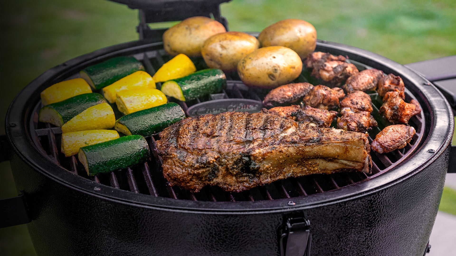 Chunks of zucchini and yellow squash grill next to whole potatoes, chicken wings, and a large steak on the grate of an AKORN Kamado grill