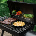 A close-up showing 3 racks of ribs and a cast iron pan of macaroni and cheese on the lower grill grate. Whole bell peppers and husk-on corn on the cob rest on the chrome plated steel warming rack.