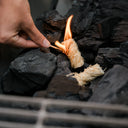 2 Char-Griller fire starters on top of a bed of lump charcoal. A person reaches in with a lit match to light the fire starters.