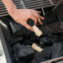 Someone places a Char-Griller fire starter on top of a pile of lump charcoal in a barrel grill