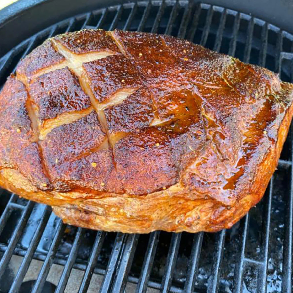 A large glazed roast with scored skin sits on the grates of a bbq grill. The drip pan is visible below the roast.