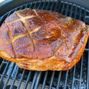 A large glazed roast with scored skin sits on the grates of a bbq grill. The drip pan is visible below the roast.