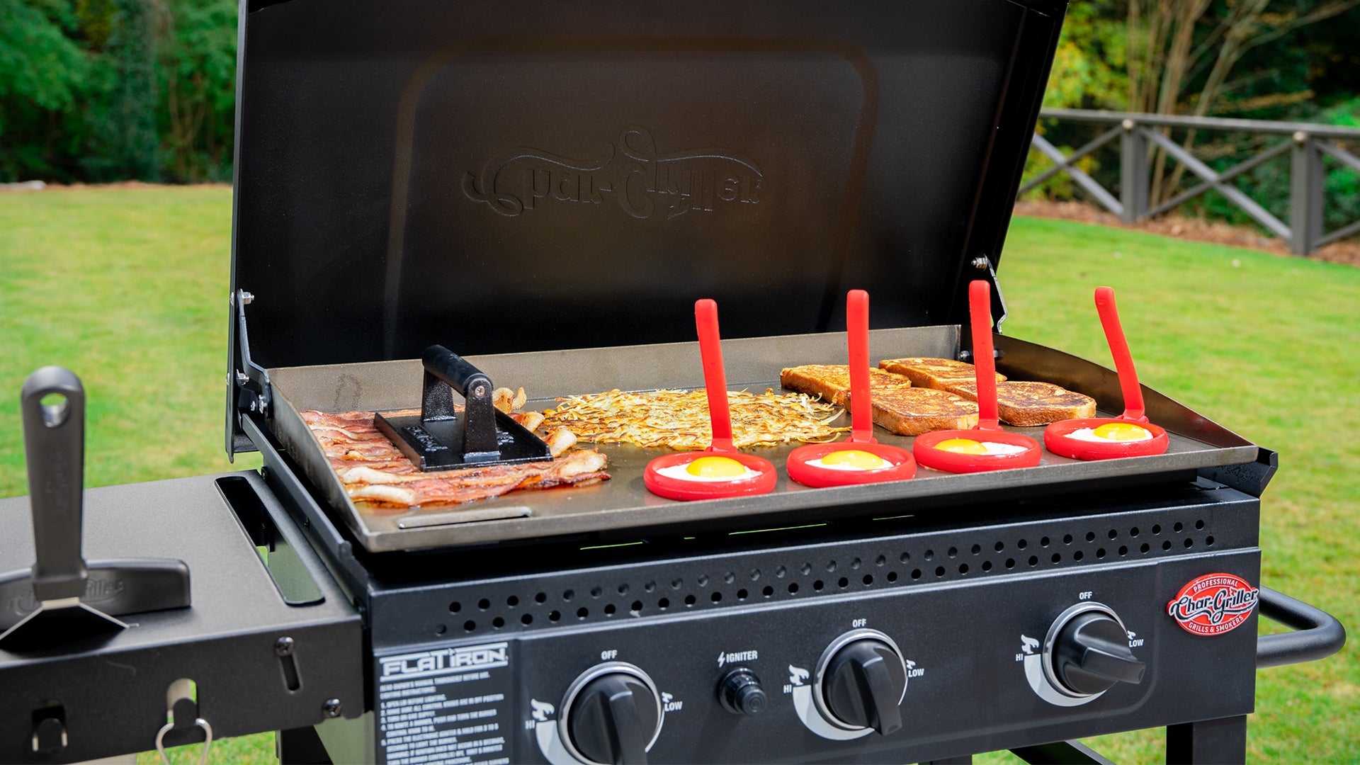 A Flat Iron griddle in use to cook bacon (using a bacon press), hash brown potatoes, French toast, and eggs (using egg rings). A wide spatula is inserted blade-down into a slot on the side shelf to the left. A green lawn and trees are in the background.