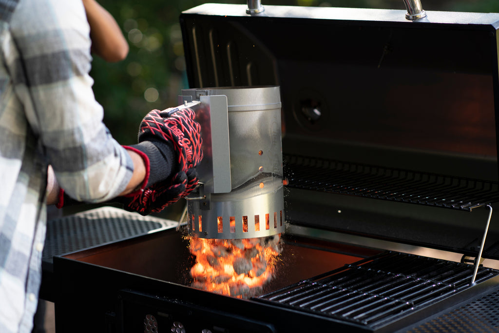 Someone wearing Char-Griller grilling gloves drops burning coals from a trigger release charcoal chimney into the bottom of a Char-Griller grill