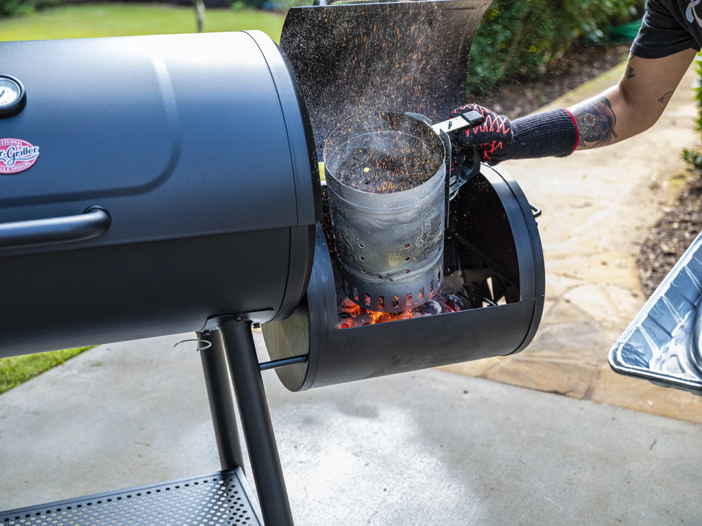 A man wearing a grilling glove uses a charcoal chimney to add burning coals to a side fire box attached to a Char-Griller barrel grill