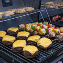 Cheeseburgers and kebabs cook on the grate of the gas side of the grill. Hamburger buns toast on the warming rack. In the background are a few sausages cooking on the charcoal side with potato wedges cooking on the warming rack above them.