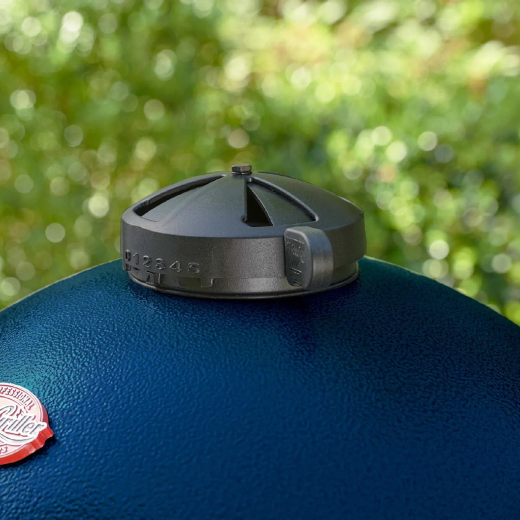 Close-up of a black top vent with embossed vent setting numbers, mounted on the blue lid of an Akorn grill with a blurred green outdoor background.