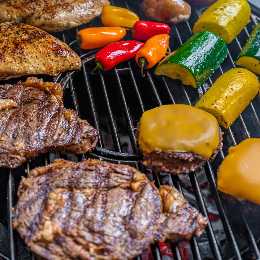 Grilled steak, chicken breast, zucchini, yellow squash, red and orange sweet peppers, and cheese-topped burgers cooking on a black grill grate.