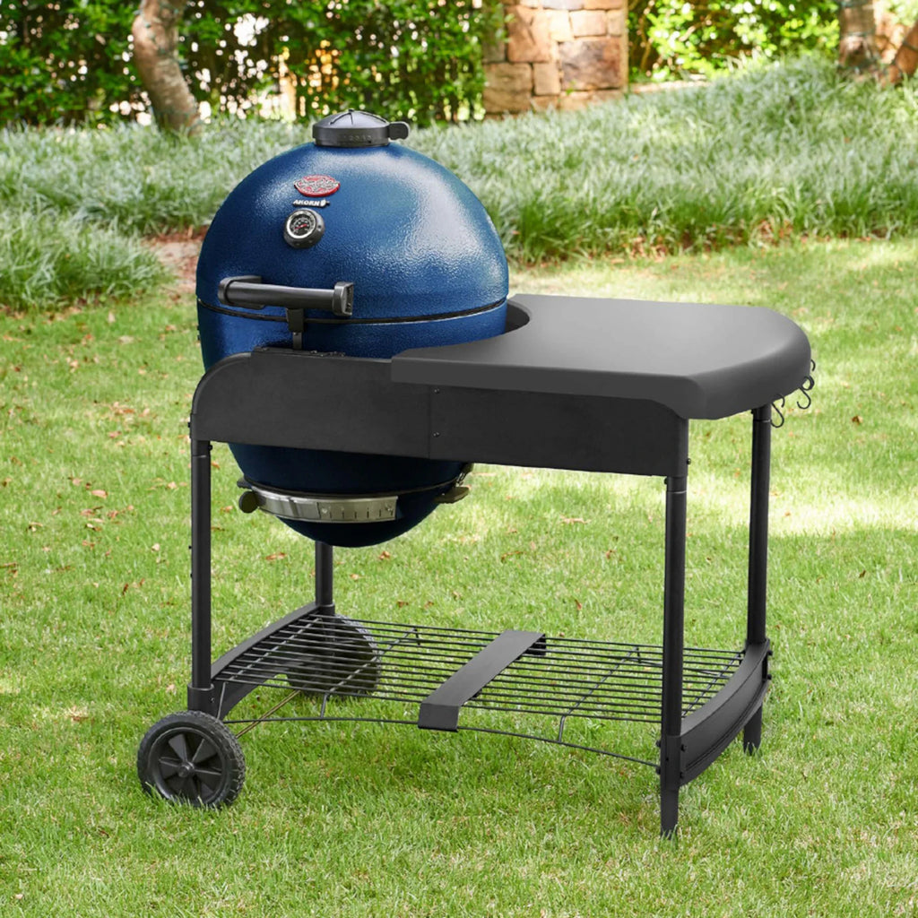 A sapphire blue kamado-style grill with a black side table and wheeled stand, photographed outdoors on a green lawn with shrubs and trees in the background. The cart is angled to show the utensil hooks mounted on the right side of the table area.