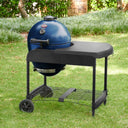 A sapphire blue kamado-style grill with a black side table and wheeled stand, photographed outdoors on a green lawn with shrubs and trees in the background. The cart is angled to show the utensil hooks mounted on the right side of the table area.