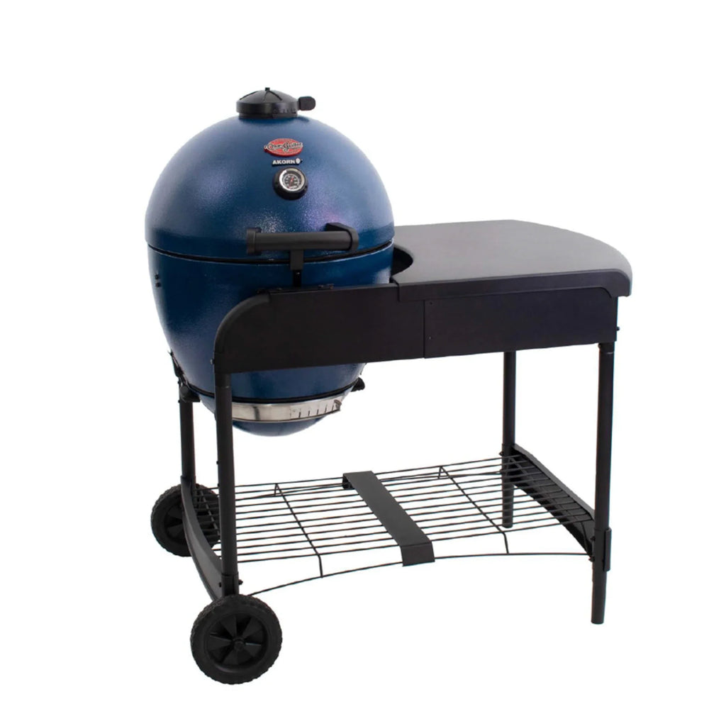 A sapphire blue Akorn kamado grill in  a cart with a black side table and wheels, shown on a solid white background.