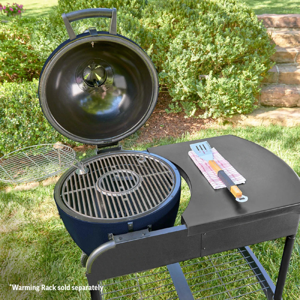 A round, blue charcoal grill with its lid open, set on a grassy lawn with shrubs in the background. The grill is mounted in the cart which has a side table, a metal grate inside, and a small warming rack on the left side of the grill. *Warming rack sold separately.
