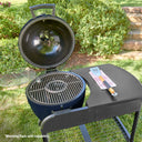 A round, blue charcoal grill with its lid open, set on a grassy lawn with shrubs in the background. The grill is mounted in the cart which has a side table, a metal grate inside, and a small warming rack on the left side of the grill. *Warming rack sold separately.