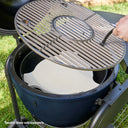 A close-up of a charcoal grill with its lid open, showing a round metal cooking grate being lifted by a hand using a grill gripper. Inside the grill is a white ceramic Smokin' Stone heat deflector.  *Smokin' Stone sold separately.