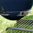 Close-up of a dark blue textured ceramic kamado with a metal draft door  and a latch holding the EasyDump Ash tray in place. Grill is positioned outdoors above the cart metal wire shelf with a blurred green grass background.