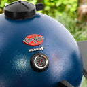 Close-up of a sapphire blue Char-Griller Akorn charcoal grill lid with a black dial thermometer and brand logo, set against a blurred green outdoor background.
