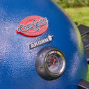 Close-up of a blue Char-Griller AKORN grill lid showing the brand badges and a built-in temperature gauge.