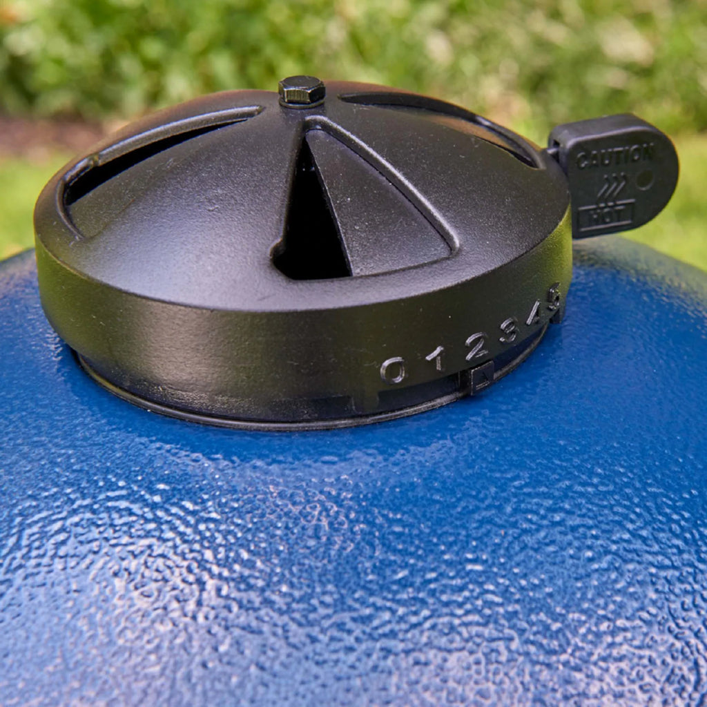 A close-up of a black top vent with adjustable slide mounted on top a sapphire blue AKORN kamado grill outdoors with blurred green foliage in the background.