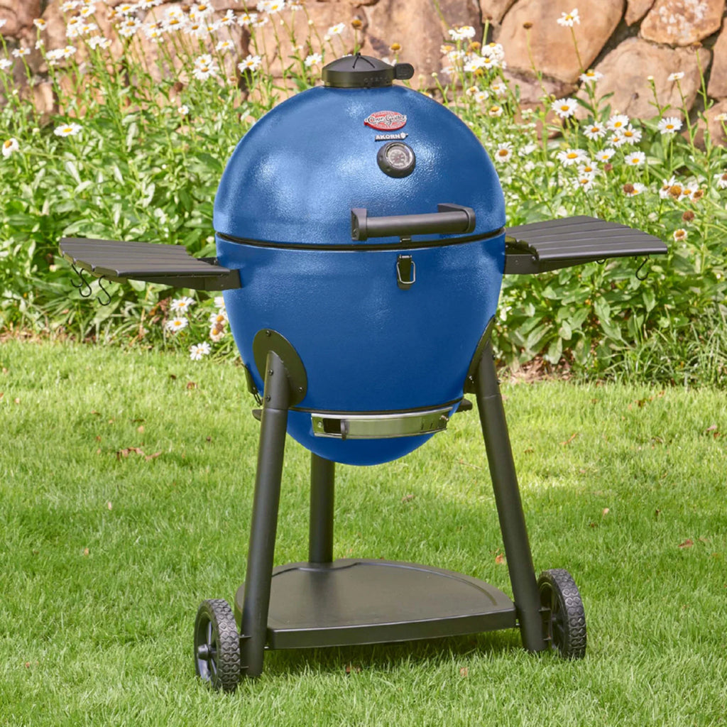 A sapphire blue kamado-style grill on a black metal cart and side shelves, photographed outdoors on green grass with flowers and a rock wall in the background.