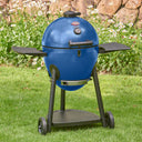 A sapphire blue kamado-style grill on a black metal cart and side shelves, photographed outdoors on green grass with flowers and a rock wall in the background.