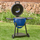 A blue kamado-style outdoor grill with the lid open and side shelves up, shown on a grassy lawn with a stone wall and garden plants in the background.