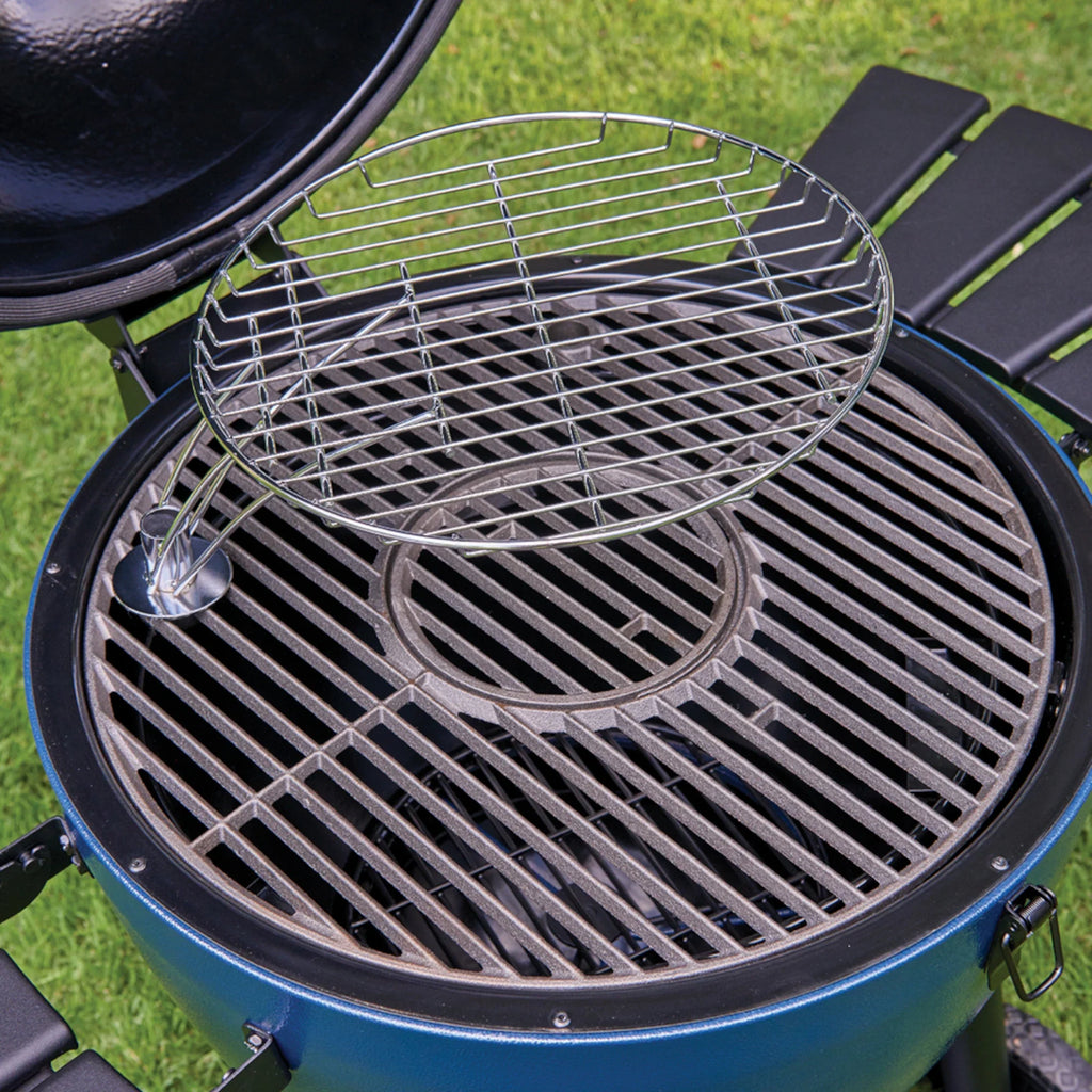 A round stainless steel wire warming rack insert resting on the inside of a blue kamado-style grill with grass visible in the background.