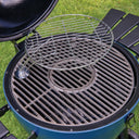 A round stainless steel wire warming rack insert resting on the inside of a blue kamado-style grill with grass visible in the background.