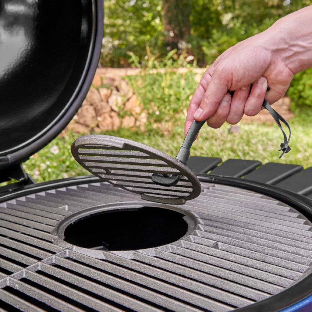 A close-up outdoor shot of a person's hand using a grate lifter to remove the round cast-iron grill grate out of the center of the primary cooking grate, with green grass and trees blurred in the background.