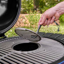 A close-up outdoor shot of a person's hand using a grate lifter to remove the round cast-iron grill grate out of the center of the primary cooking grate, with green grass and trees blurred in the background.