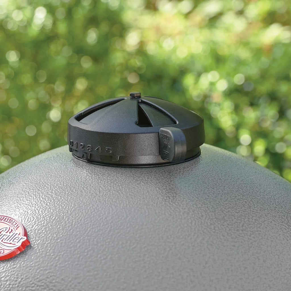 Close-up of a black top vent with embossed vent setting numbers, mounted on the grey lid of an Akorn grill with a blurred green outdoor background.