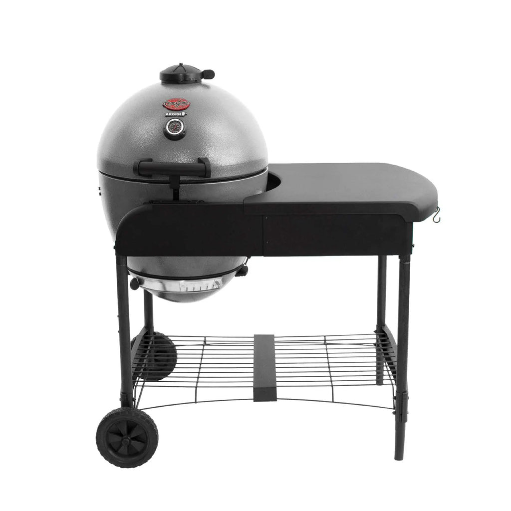 A ash grey Akorn kamado grill in a cart that has a black side table, 2 wheels on the left side under the grill, and a wire shelf between the cart legs. Shown on a solid white background.
