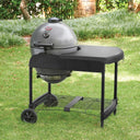 A ash grey kamado-style grill with a black side table and wheeled stand, photographed outdoors on a green lawn with shrubs and trees in the background. The cart is angled to show the utensil hooks mounted on the right side of the table area.