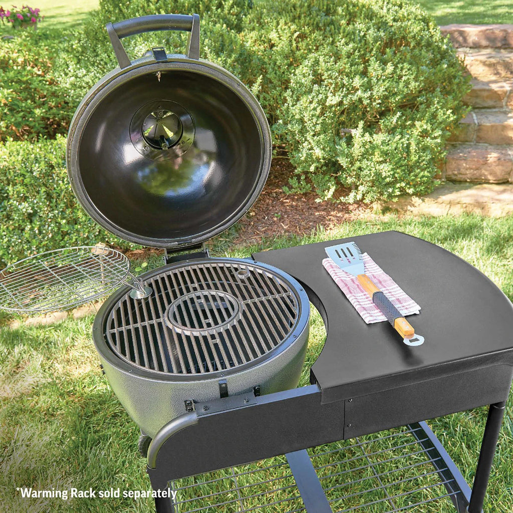 A round, grey charcoal grill with its lid open, set on a grassy lawn with shrubs in the background. The grill is mounted in the cart which has a side table, a metal grate inside, and a small warming rack on the left side of the grill. *Warming rack sold separately.